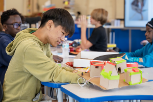 A close-up photo of a young student working with his teammates to construct a mini house.