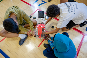 Three students sit on the floor of a gym as they work together to replace the wheels on a miniature race car.