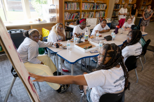 A group of young female students are seated at a table while program administrators look on in the background. A young female student points to the presentation board as she speaks on behalf of her team members during the student showcase.