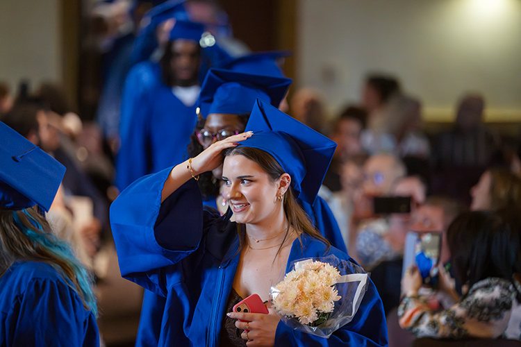 A female graduate of the adult education program holds her cap as she walks down the aisle during the ceremony. Several other graduates are walking behind her.