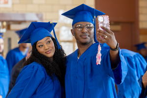 A female and male student wearing royal cap and gowns pose for a selfie together at the adult education graduation ceremony.