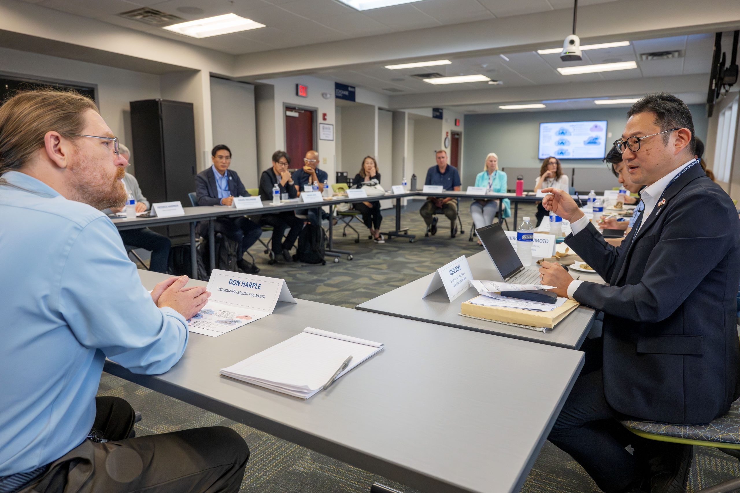 A group of people sit in a conference room, the tables are arranged in a square. In the foreground, two people are speaking to each other. One is gesturing, the other is clearly listening