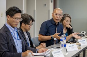 A man is sitting at a conference room table with other people. He is speaking and gesturing