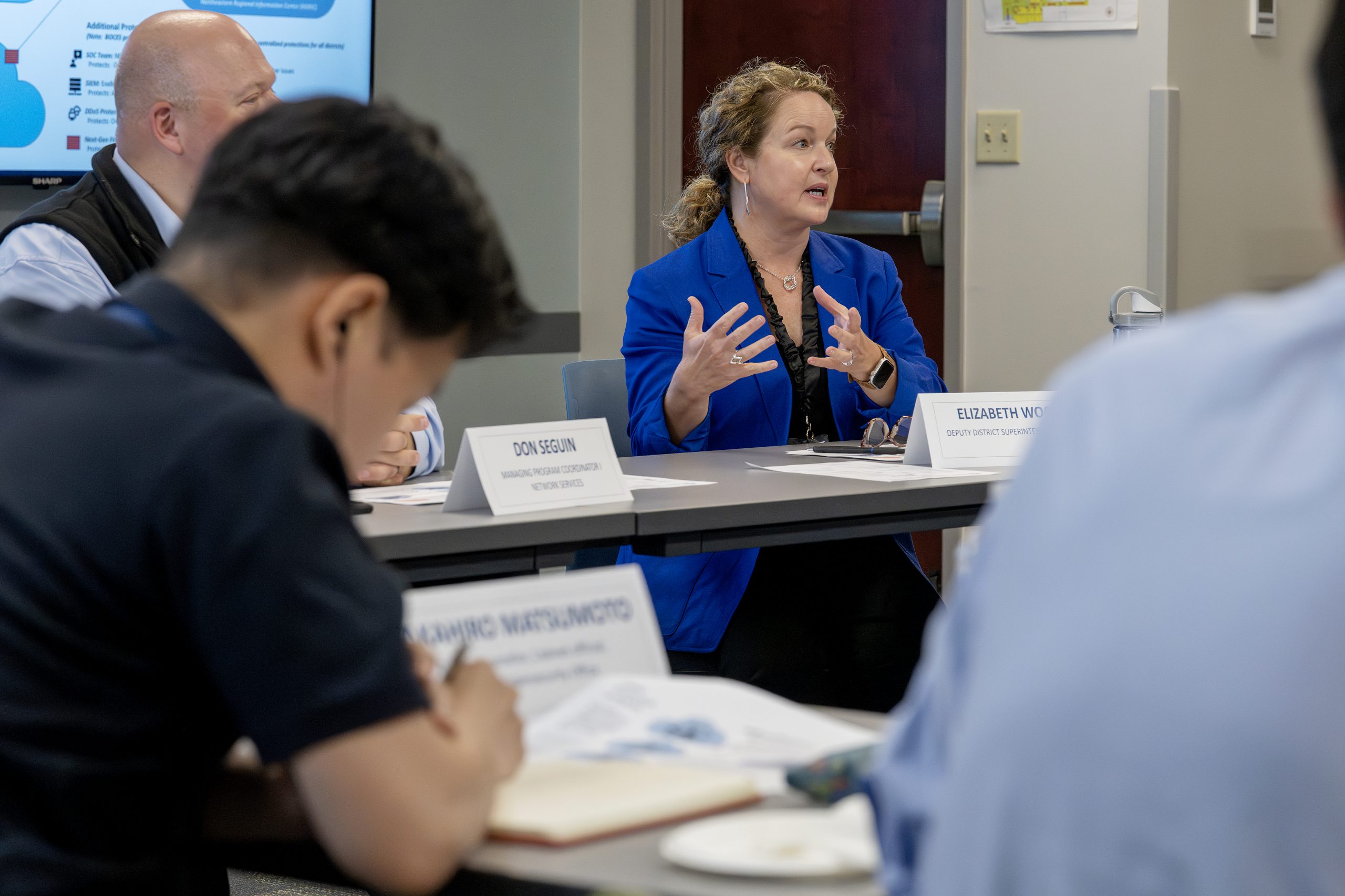 People are sitting at conference room tables. A woman is in the center of the photo, she is wearing a bright blue blazer and is speaking and gesturing