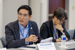 Two people are sitting at a conference room table. One is looking down at papers and the other is speaking to someone off camera