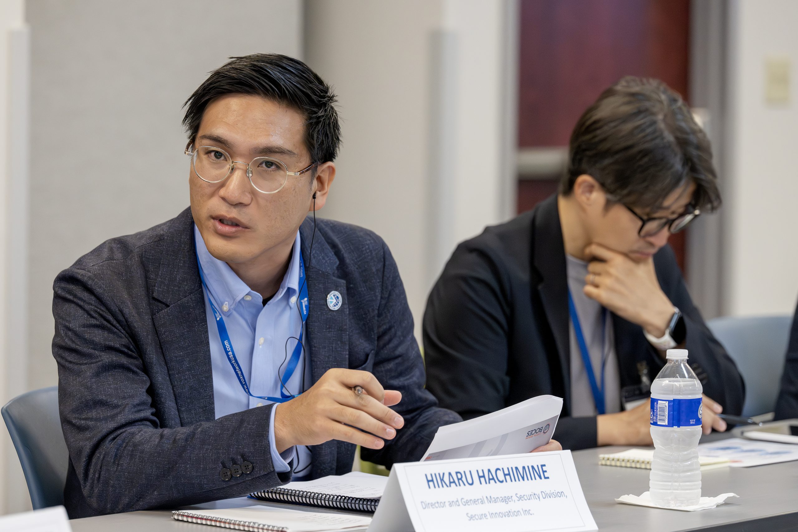 Two people are sitting at a conference room table. One is looking down at papers and the other is speaking to someone off camera