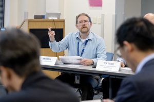 In a conference room, a man is speaking and gesturing. In the foreground, blurred out a bit, you can see the back of the heads of two other people