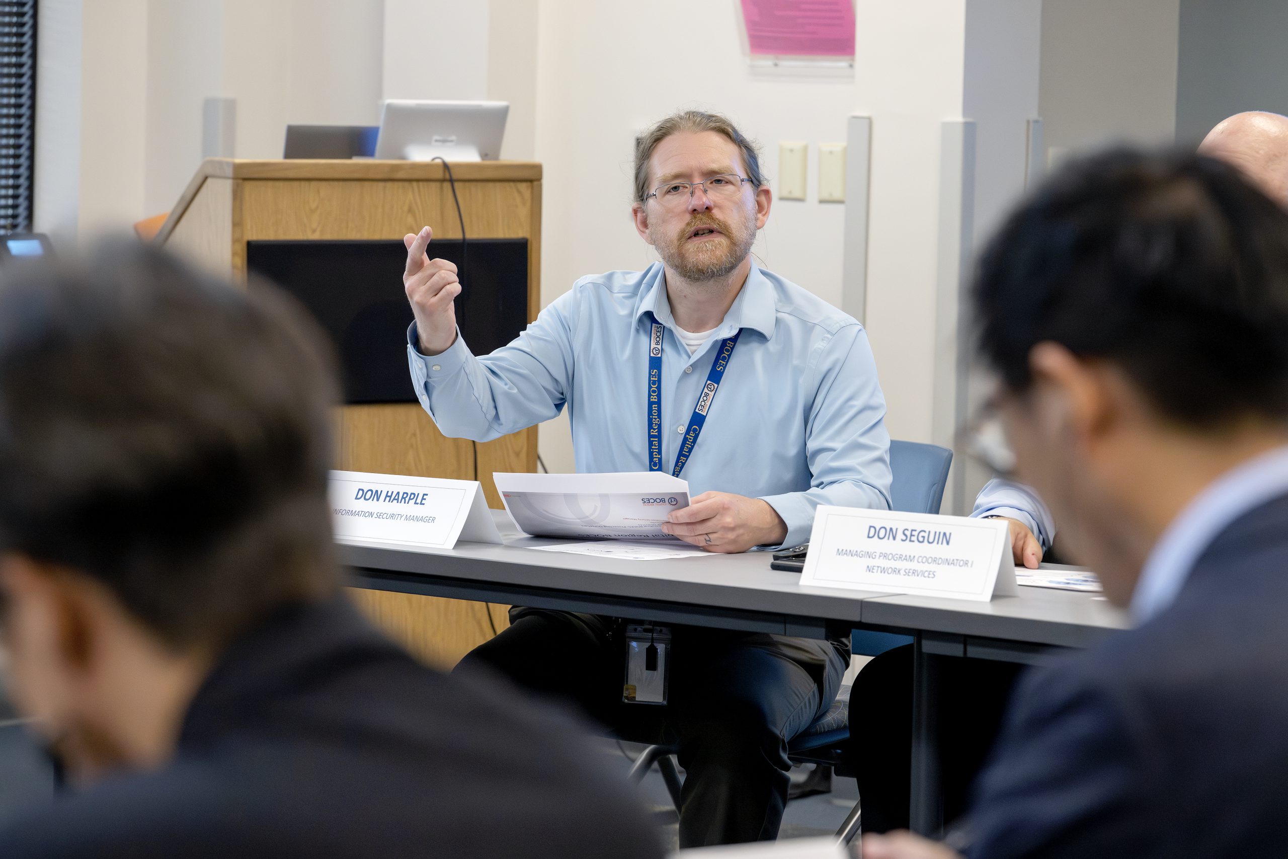 In a conference room, a man is speaking and gesturing. In the foreground, blurred out a bit, you can see the back of the heads of two other people
