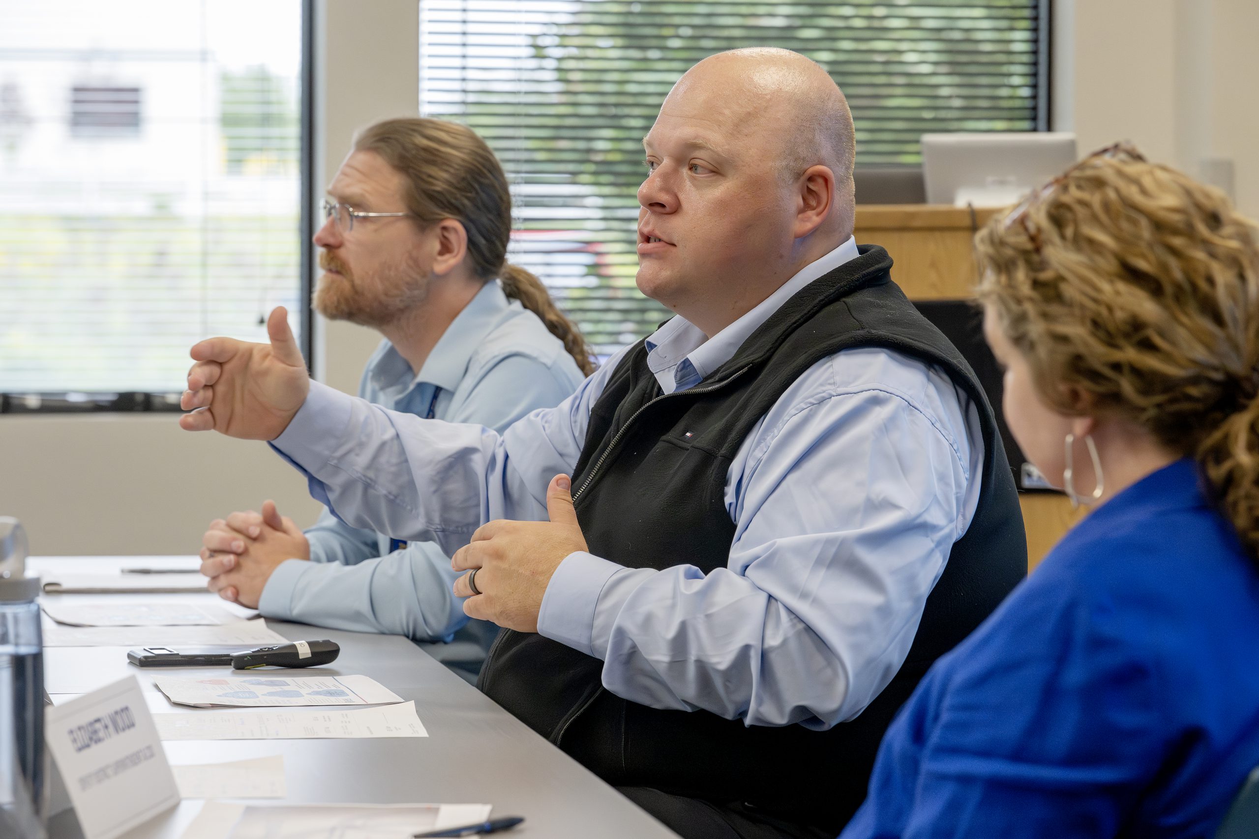 In a conference room, three people are sitting at a table, one is speaking and gesturing