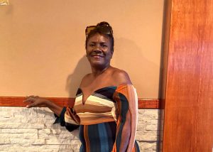 Yvonne posses in a jump suit in front of a brown wall