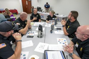 A group of police officers are seated at a conference room table. One is speaking