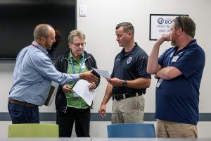 Four people are discussing something. One man is holding a piece of paper, one is gesturing