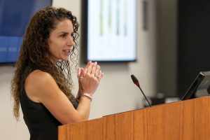 A woman stands at a podium speaking to an audience. There's a monitor behind her