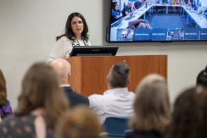 A woman stands at a podium speaking to an audience. There's a monitor behind her