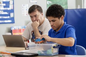 An Occupational Therapy Assistant is photographed  looking into a laptop as he sits next to a student who is working with blue puddy. 