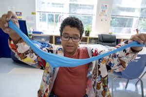 A young student is photographed in his classroom as he uses both arms to stretch blue puddy.