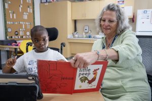 A teacher at Maywood Academy hold a book open as she and a young male student look into the book. 
