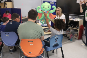 Students listen intently as the author reads from the book. A young student joyfully raises his Ocho the Octopus toy.
