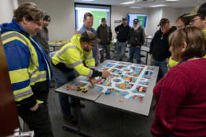 A group of people are standing around a table, many are wearing clothing that is bright yellow. On the table is a child's map of a town with toy trucks on the table as well. One person is using the trucks in an exercise to show traffic flow.