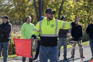 A participant in a flagger training is wearing a bright green/yellow and black sweatshirt. He's holding an orange flag in his right hand and has his left hand raised, signifying stop.