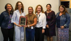 Six people are standing in a row, facing the camera. One is being presented with a plaque